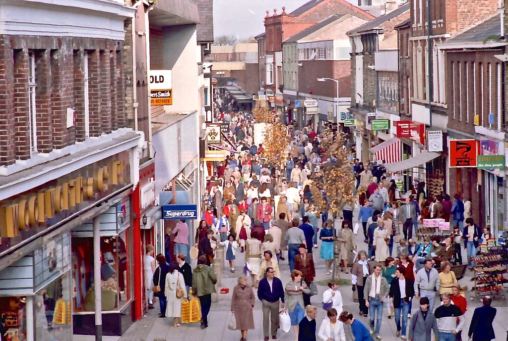 A crowded pedestrian high street on a Saturday in 1990, with Woolworths and Superdrug on the left and shoppers carrying bags